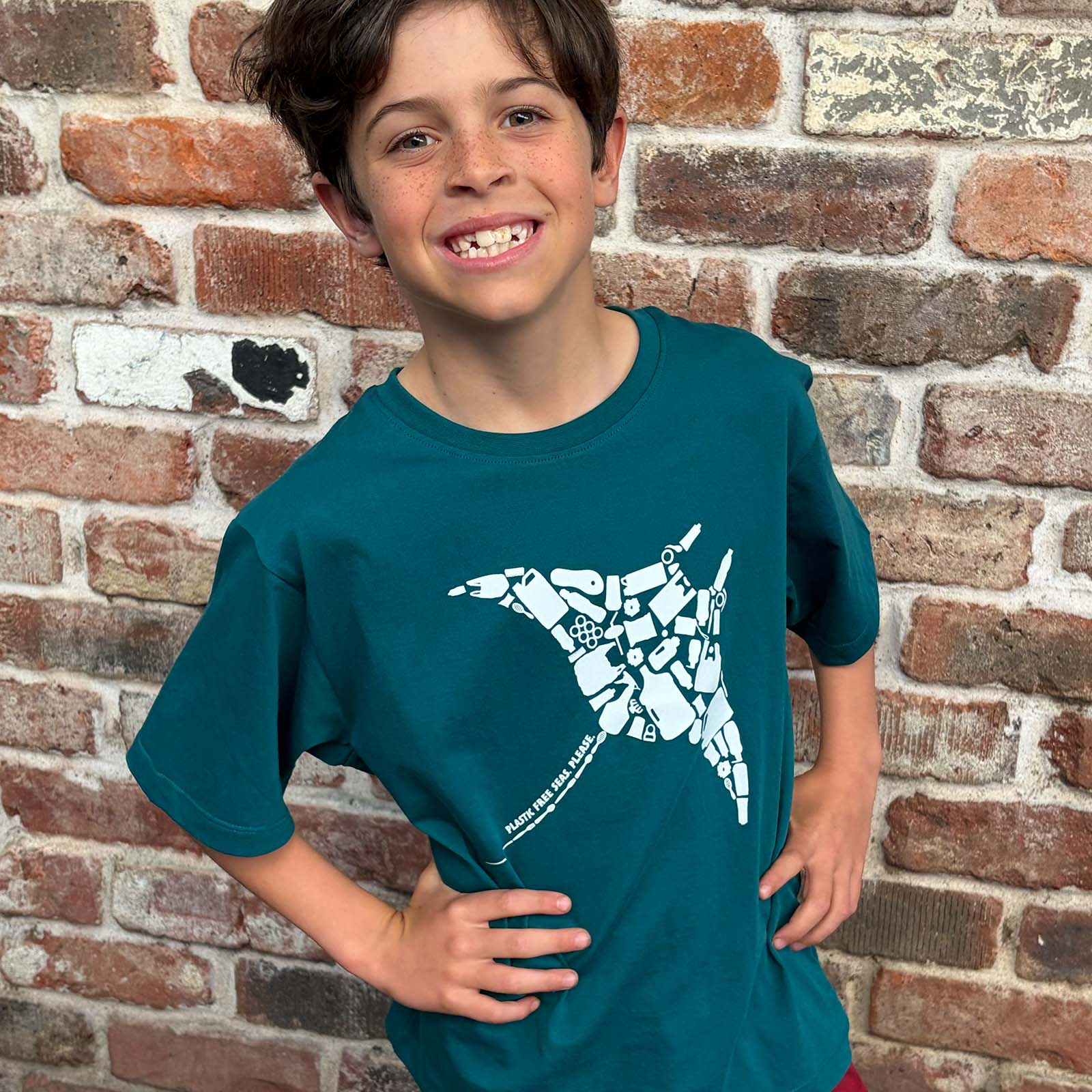 A smiling boy wearing a South Beach Boardies Seagreen t-shirt with a white manta ray plastic free seas please graphic, standing in front of a recycled brick wall.