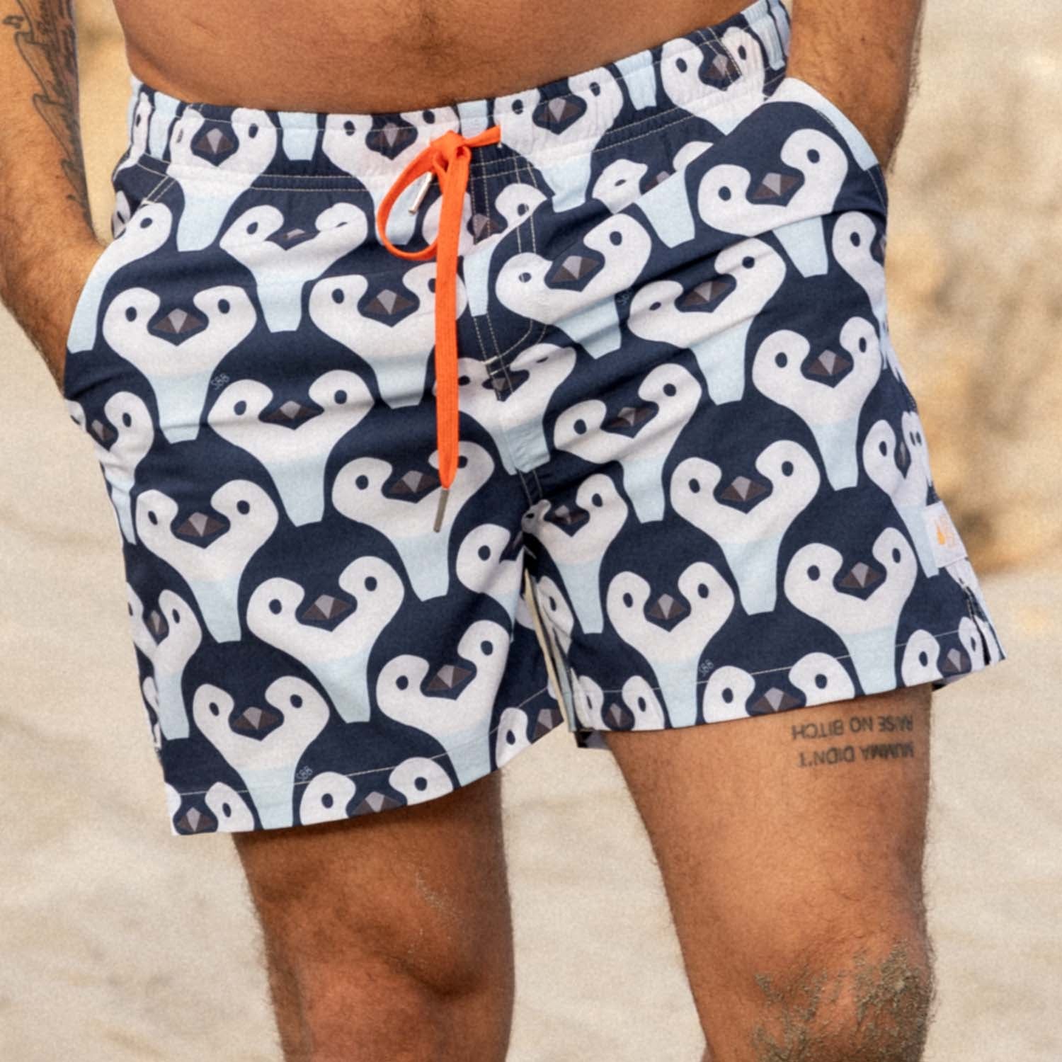Person wearing swim shorts with white penguin pattern at an Australian beach. The board shorts are Men's Stretchy Trunks in Penguin Parade print made from Recycled Plastic Bottles by South Beach Boardies