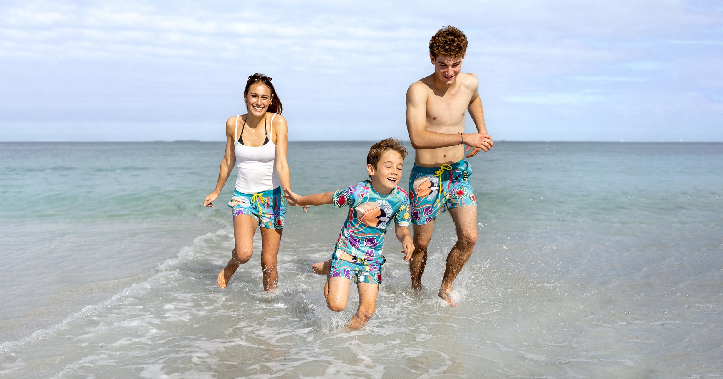 Family of three running on a beach with ocean and sky in the background, SOUTH BEACH BOARDIES MADE FROM RECYCLED PLASTIC BOTTLES MATCHING LIMITED EDITION PRINTS FOR FAMILIES , IN COLOURFUL HELEN ANSELL AUSTRALIAN PRINT