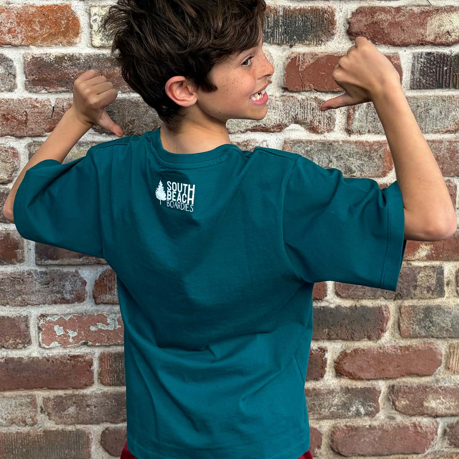 A boy wearing an organic cotton Seagreen short-sleeve t-shirt with a white 'South Beach Boardies' logo on the back, posing with his hands near his head in front of a brick wall.