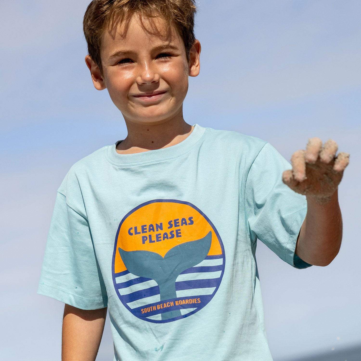 Child wearing a light blue t-shirt with a whale graphic and text against a clear sky in Fremantle, Kids Organic Cotton Tshirts and UPF50+ Recycled Rashies by South Beach Boardies.psd