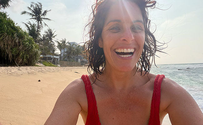 Kirsten Lopez in a red swimsuit on a beach with palm trees and ocean in the background in Sri Lanka. Kirsten Lopez is the founder of South Beach Boardies Better for the Planet Beachwear.