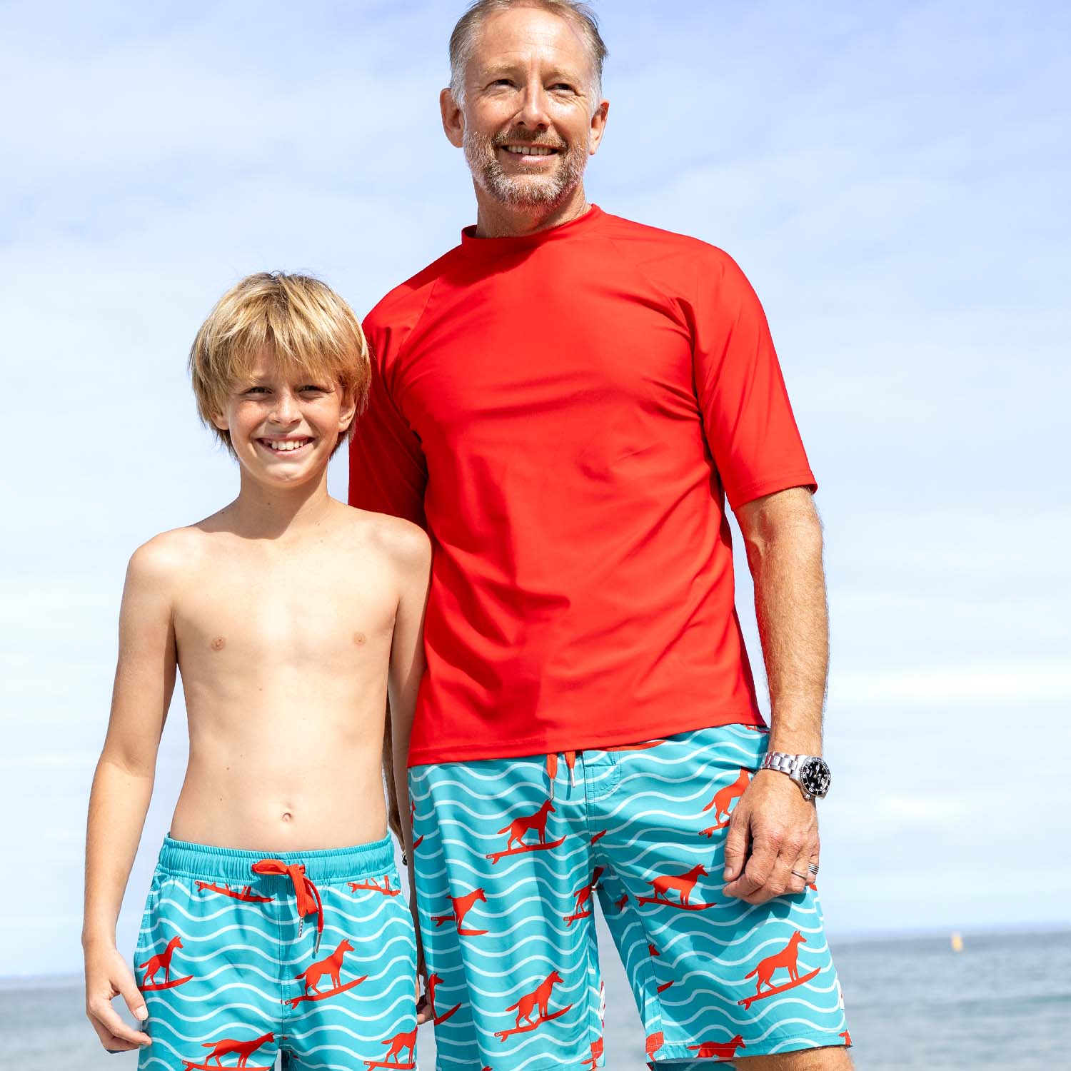 Man and son wearing matching blue swim trunks with red surfing dingo designs on a Fremantle beach, part of the Mens Recycled UPF50+ Rashies and Organic Cotton Tshirts from South Beach Boardies