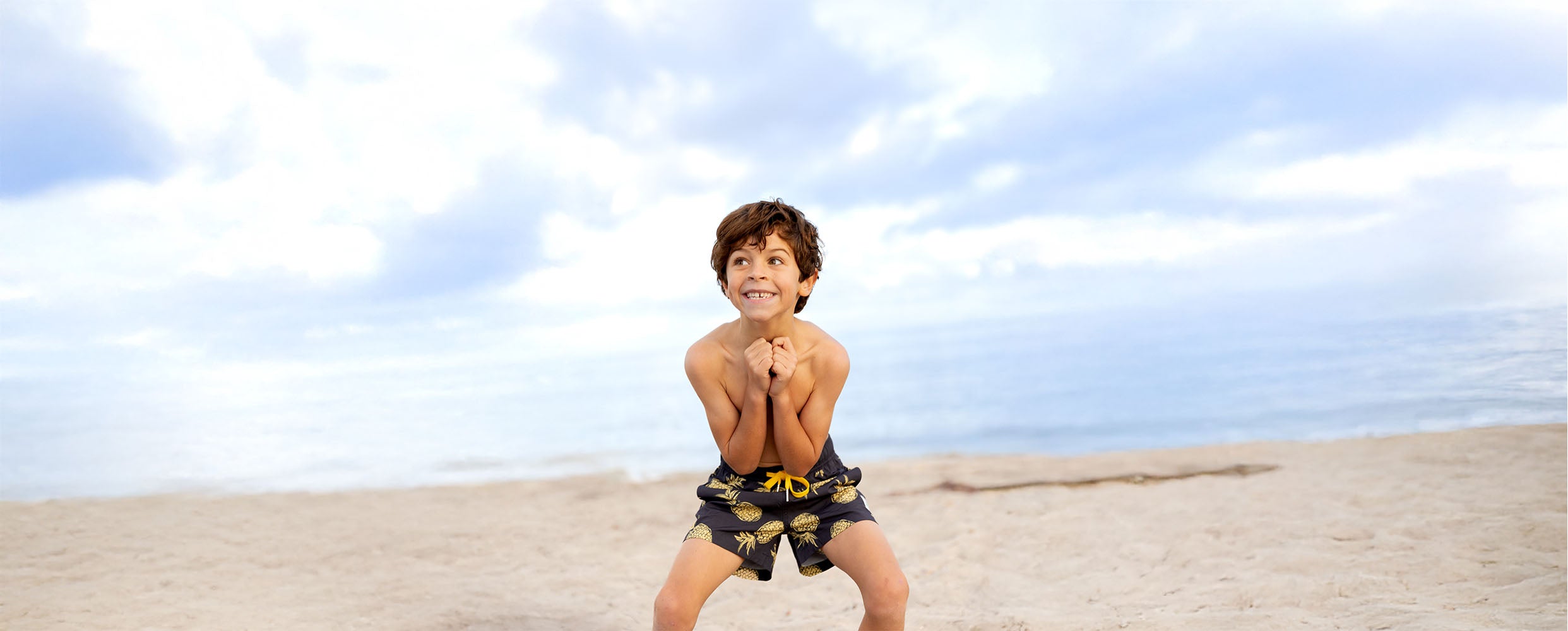 Child in swim trunks posing on a sandy West Australian beach with a blue sky. Kids Stretchy Trunks collection Gold Pineapples by South Beach Boardies.