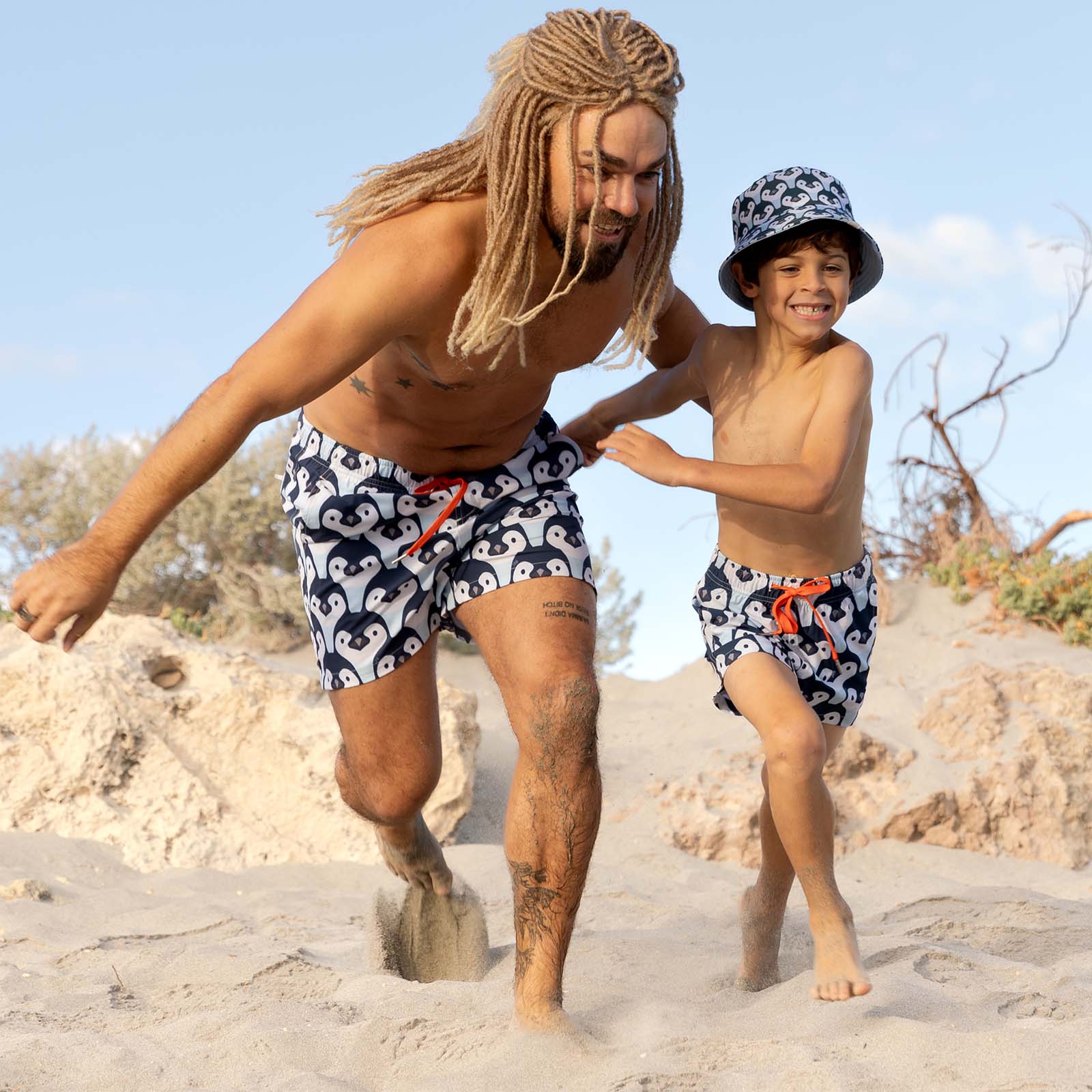Man and child playing on a sandy beach wearing matching Penguin swim trunks. These are South Beach Boardies best-selling family matching swimwear made from Recycled Plastic Bottles