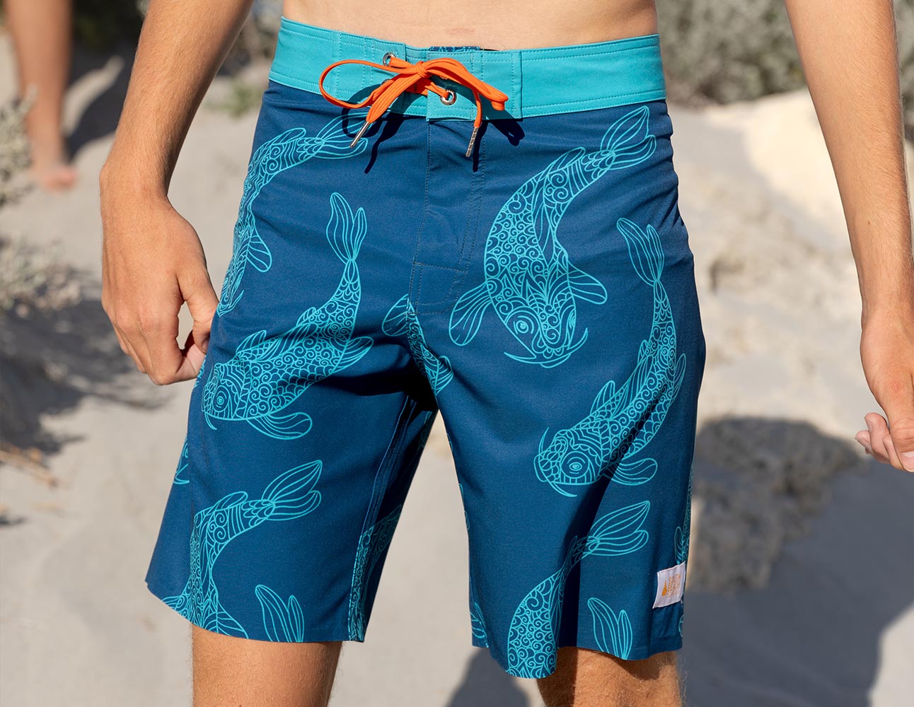 Man wears Blue swim shorts with Koi carp pattern worn on an Australian beach. These shorts are South Beach Boardies Mens Technical Performance Boardies Made from Recycled Plastic Bottles