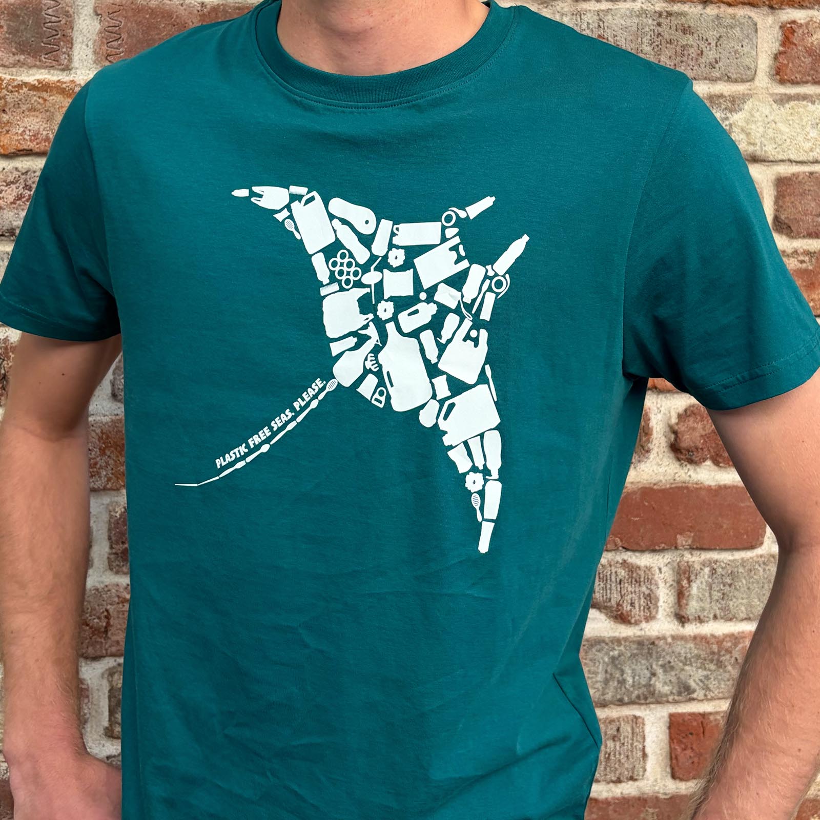 Man wears seagreen Unisex Organic Cotton Tshirt Rays Plastic Free Seas Please, by South Beach Boardies, standing against brick wall