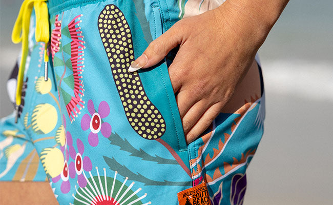 Colorful floral-patterned shorts with a hand adjusting the drawstring. THESE ARE WOMENS STRETCHY BOARD SHORTS WITH POCKETS MADE FROM RECYCLED PLASTIC BOTTLES BY SOUTH BEACH BOARDIES