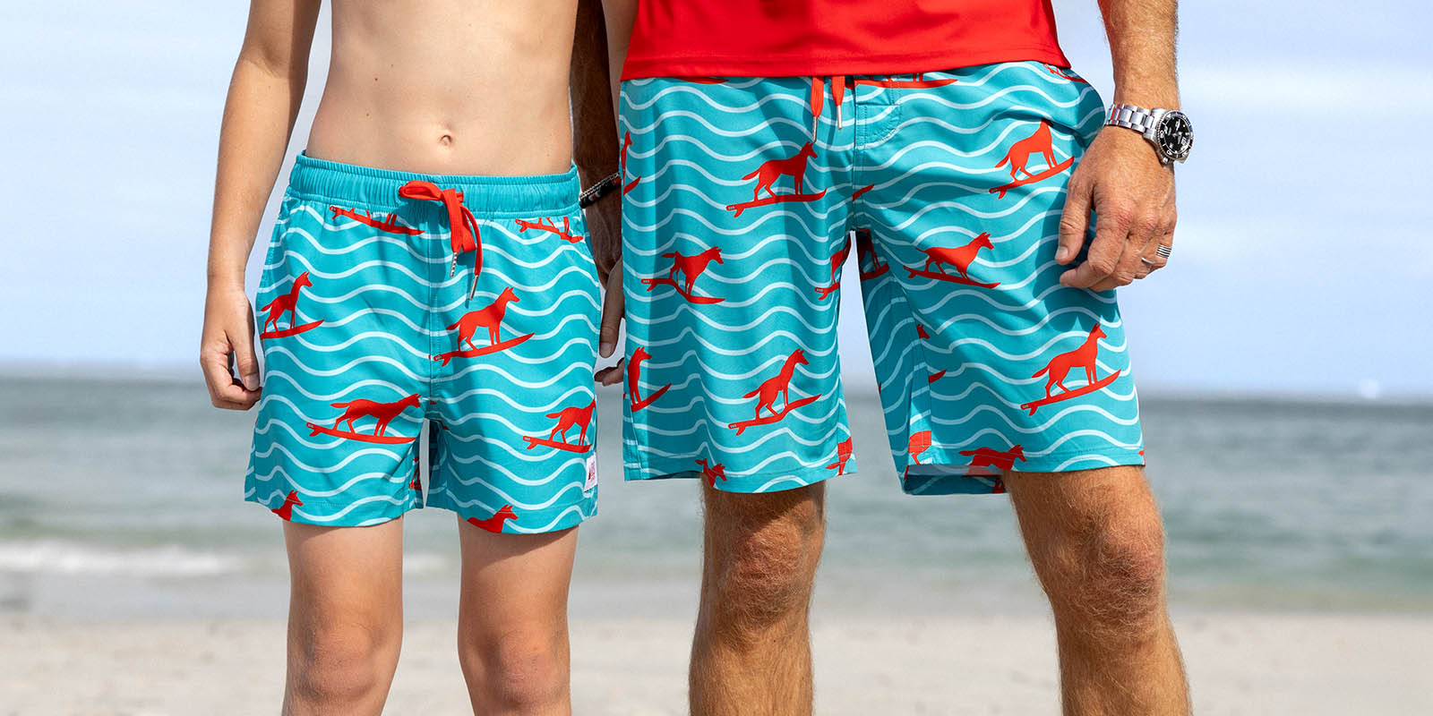 Two people wearing blue swim shorts with red dingo patterns on a beach. matching truquoise surfing dingo boar=dies made from recycled plastic bottles by su=outh beach boardies.